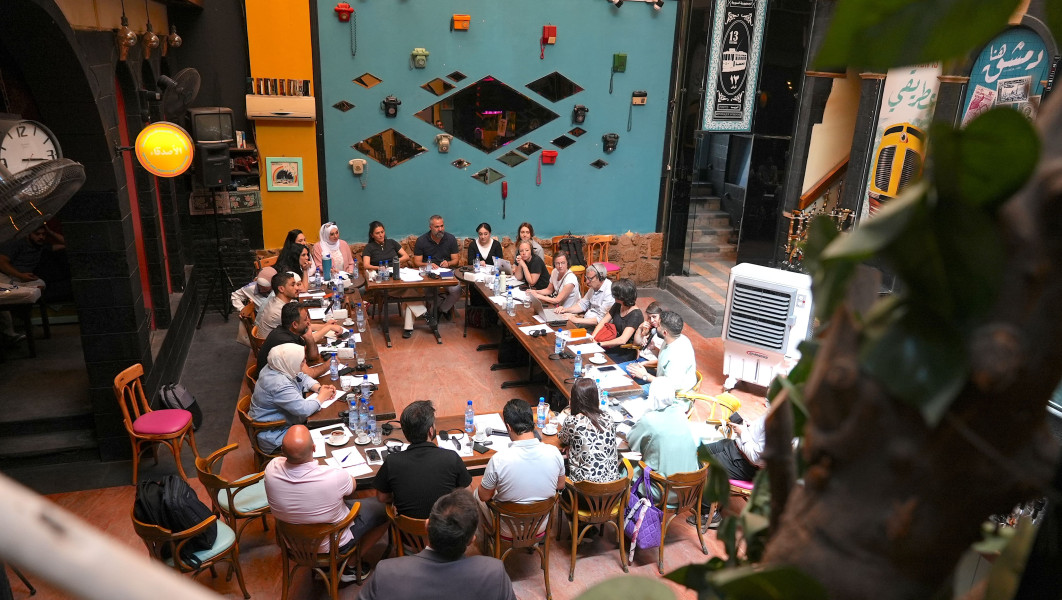Approximately 25 people are seated around several long tables configured in a large rectangle in a big room.
