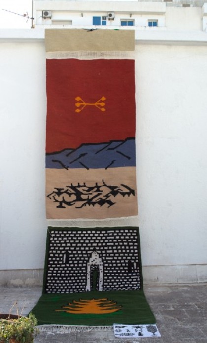 A red, blue, tan, black and green tapestry hangs on an outdoor wall, over a cobblestone street.