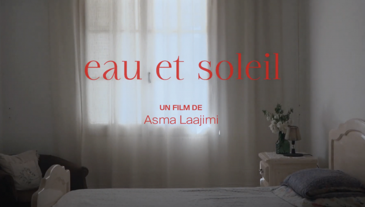 A still image pulled from a short film, of a bed room with curtains pulled, with the French words, "Eau et soleil, un film de Asma Laajimi."