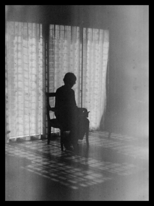 Black and white photograph of a figure sitting in a chair in the center of a room, with light coming through curtains behind them.